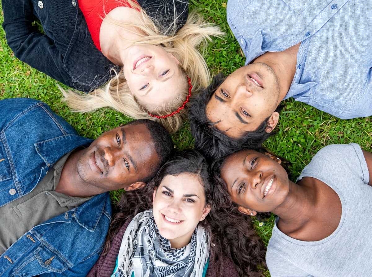 Five students lying on the grass in a circle, heads together, smiling up at the camera.
