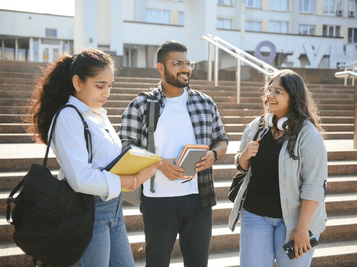 Three students talking together on campus steps in bright sunshine, folders and tablets in hand.