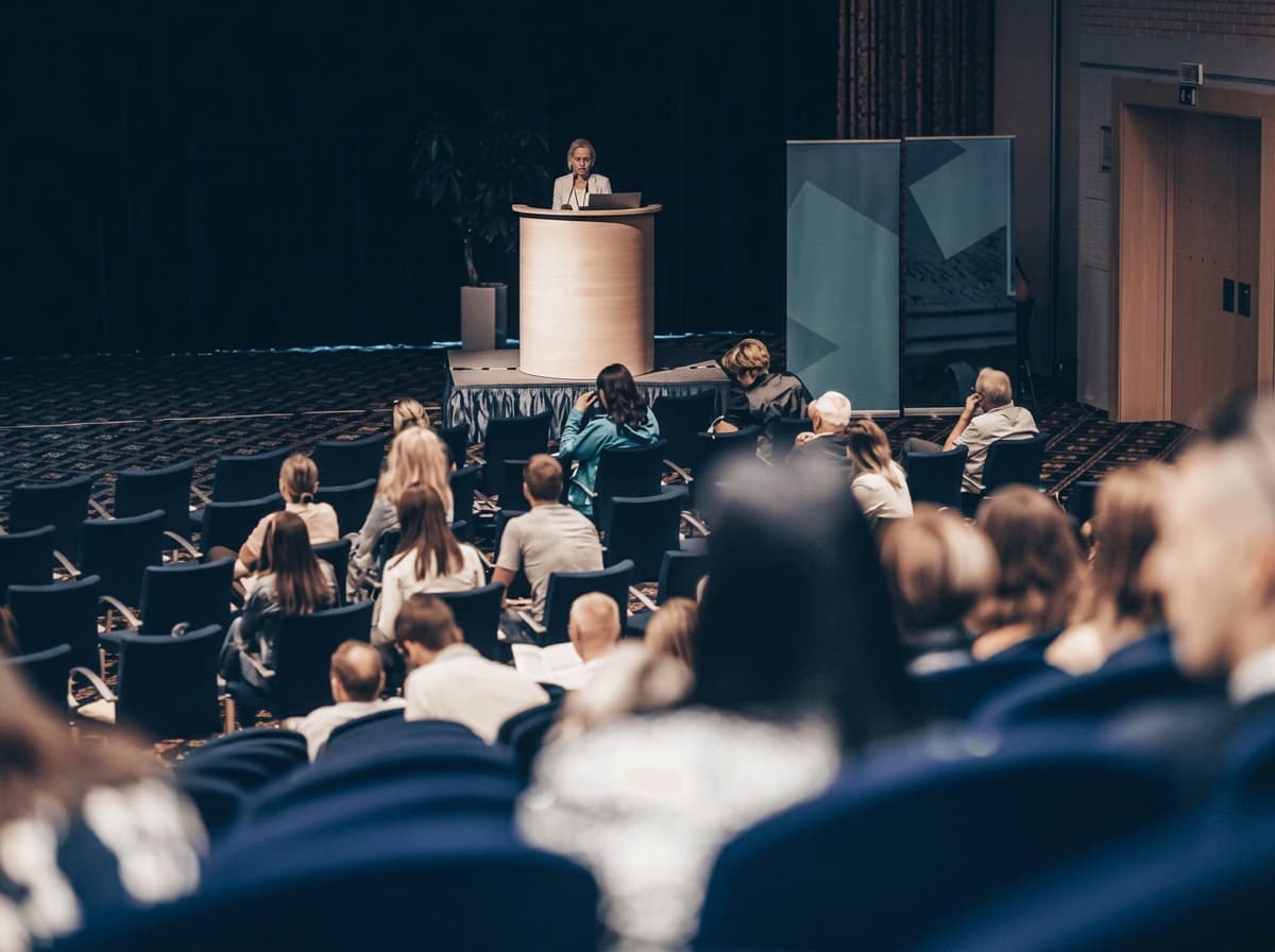 A speaker presenting at a lectern in a darkened conference hall.