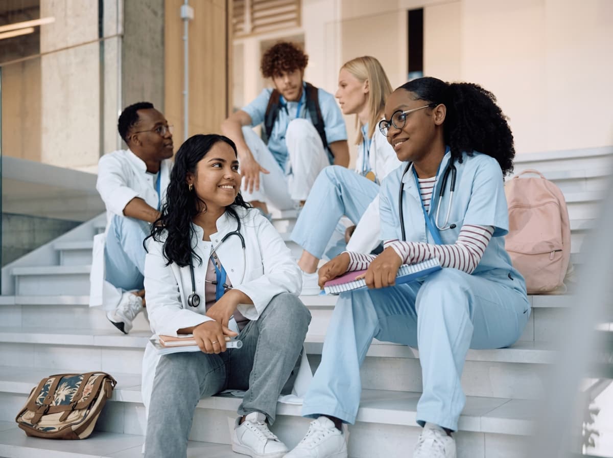 Healthcare students in scrubs sitting and talking together on campus steps.