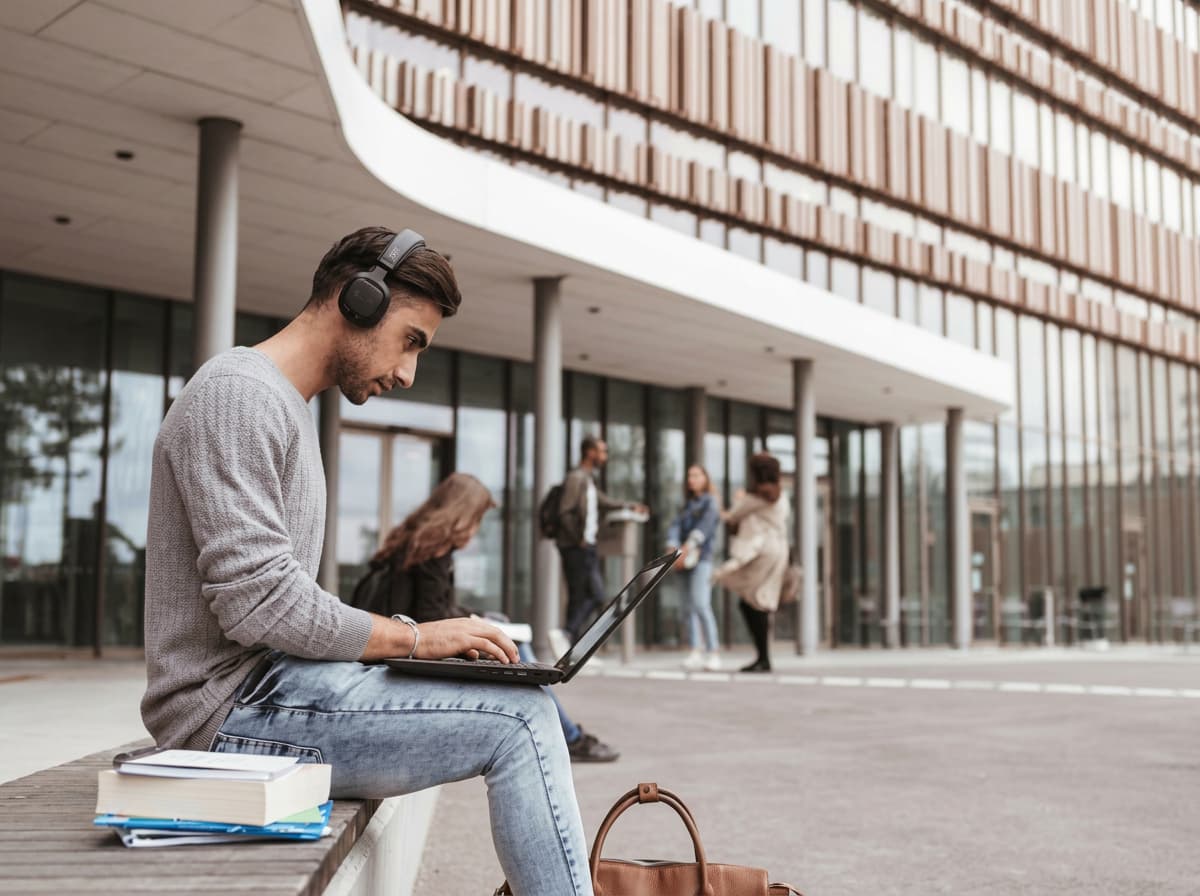 A student working on a laptop on a bench outside a modern louvred building.