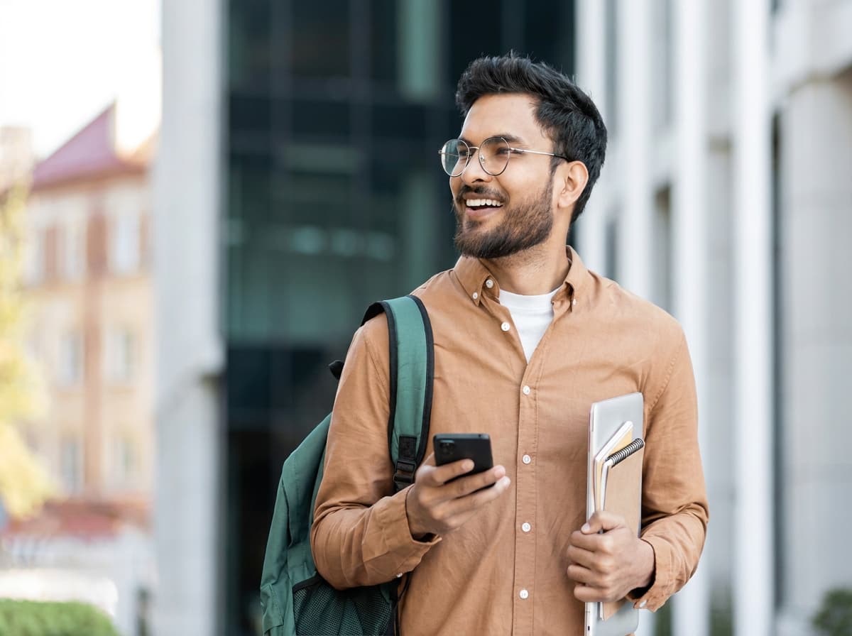 A student carrying notebooks and a backpack outside a modern glass university building.