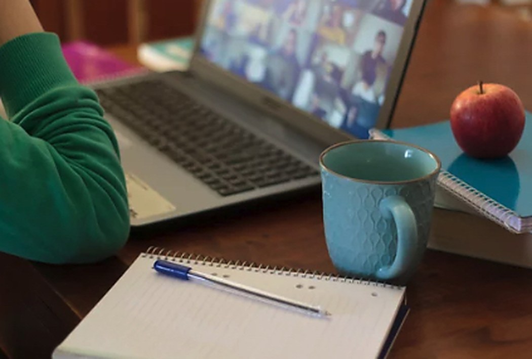 A laptop showing a video-call grid beside a notebook and mug on a home desk.