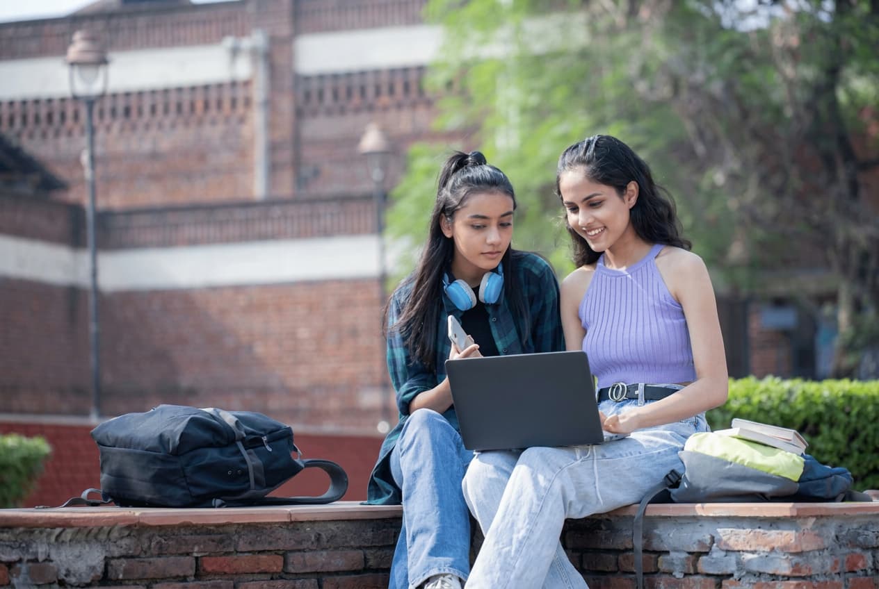 Two students from India studying together on a campus wall with laptops.