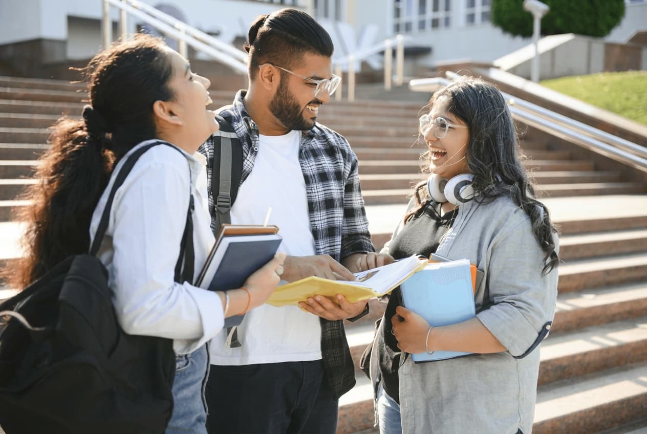 Three students talking and laughing together on sunlit campus steps.