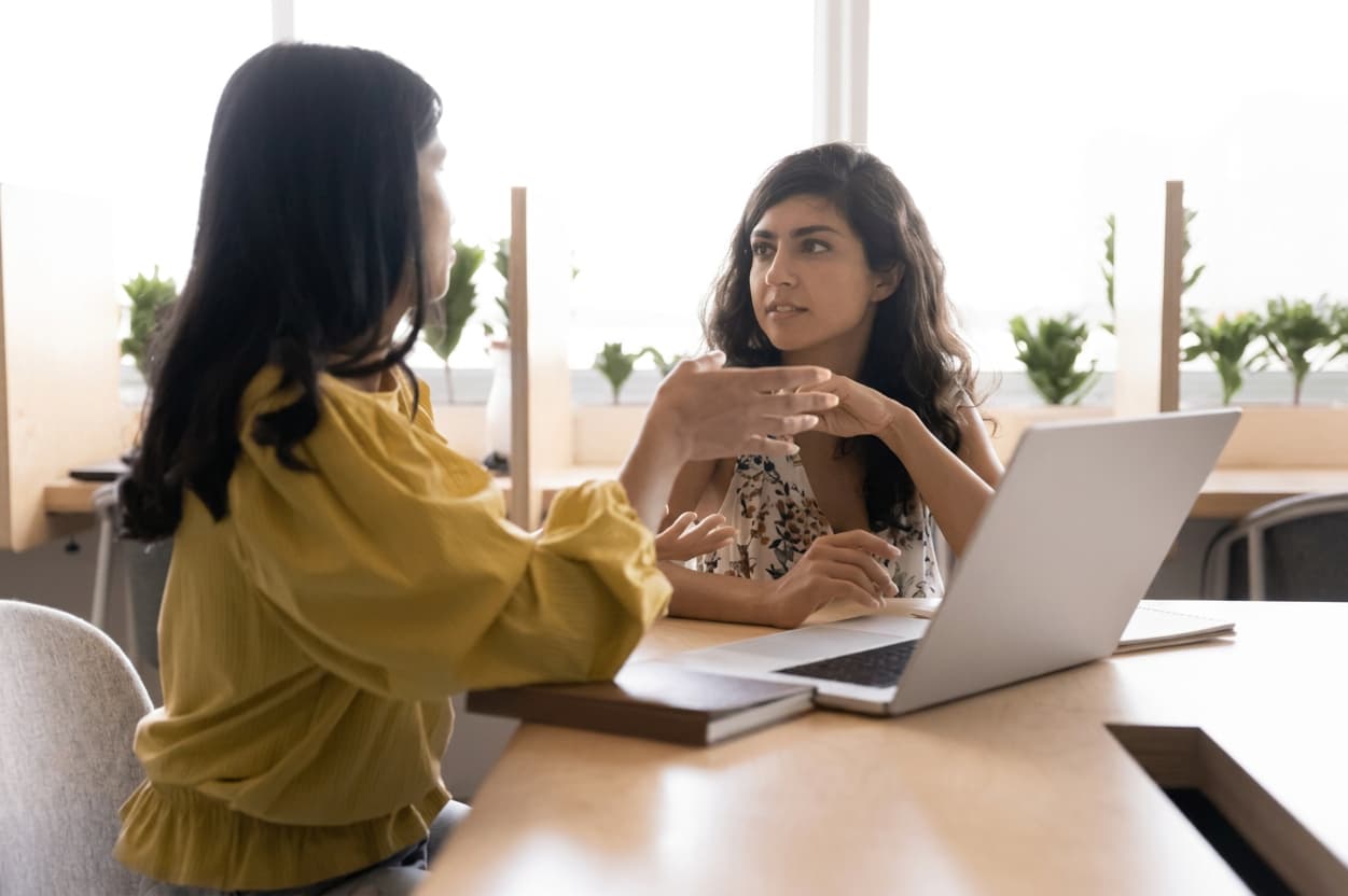 Two students talking across a light wooden table beside an open laptop.