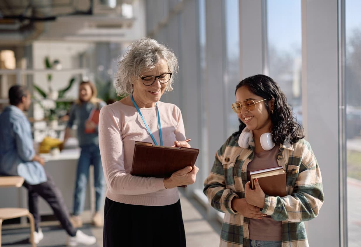 An adviser writing on a clipboard beside a smiling student holding books in a bright campus atrium.