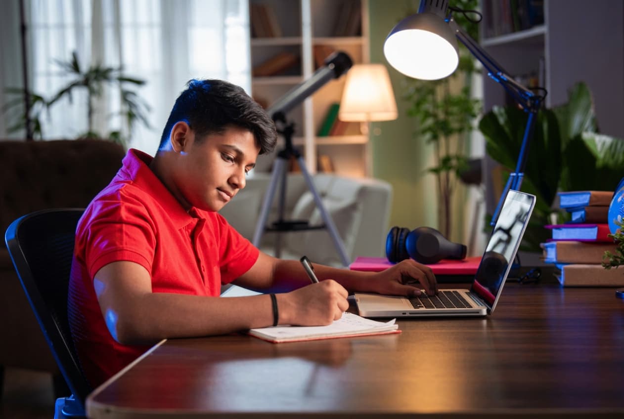 A student writing in a notebook beside an open laptop at a desk lit by a single lamp at night.