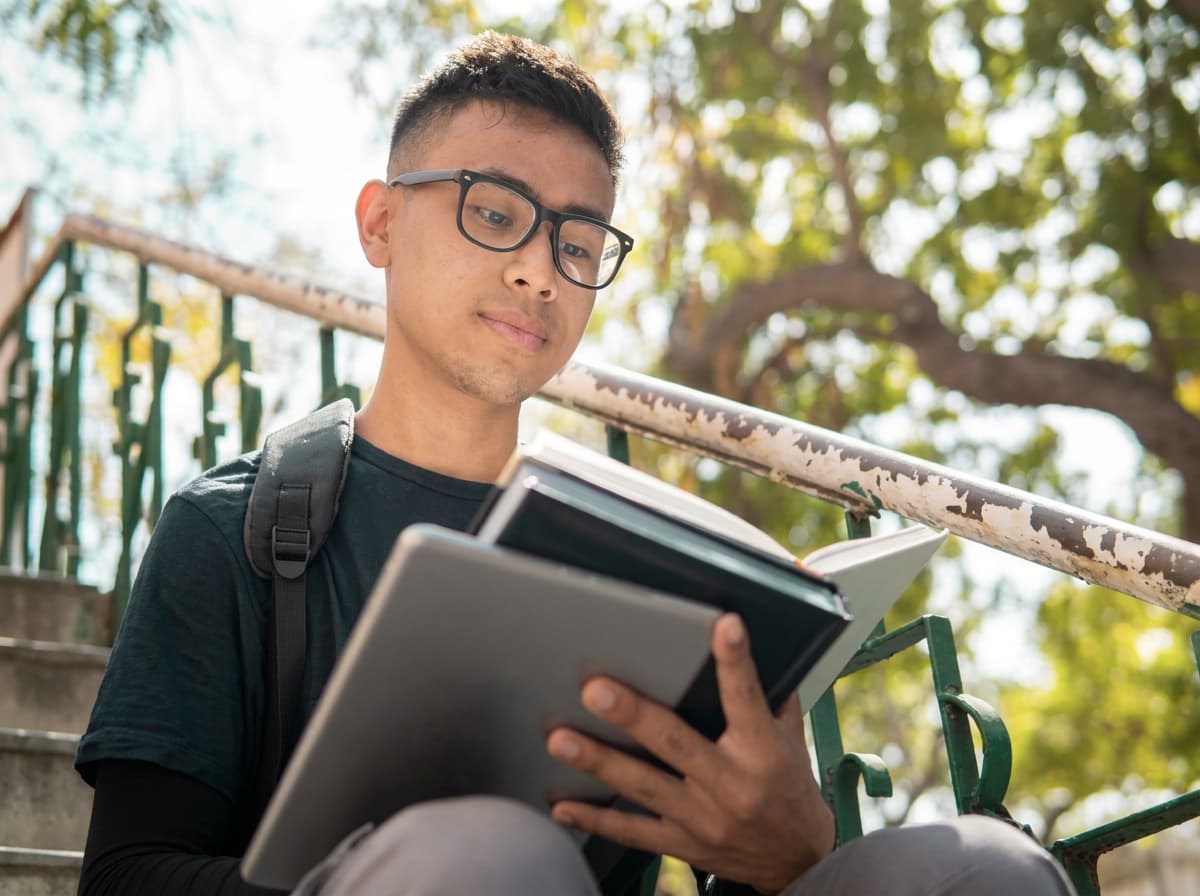 A student reading a book and holding a tablet on sunlit campus steps.