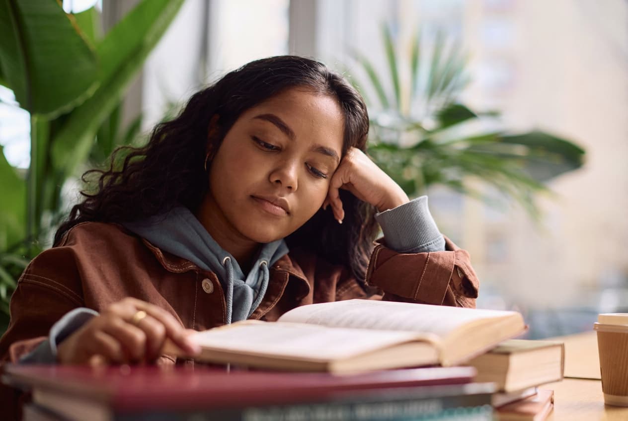 A student reading at a sunlit desk with a book stack and coffee.