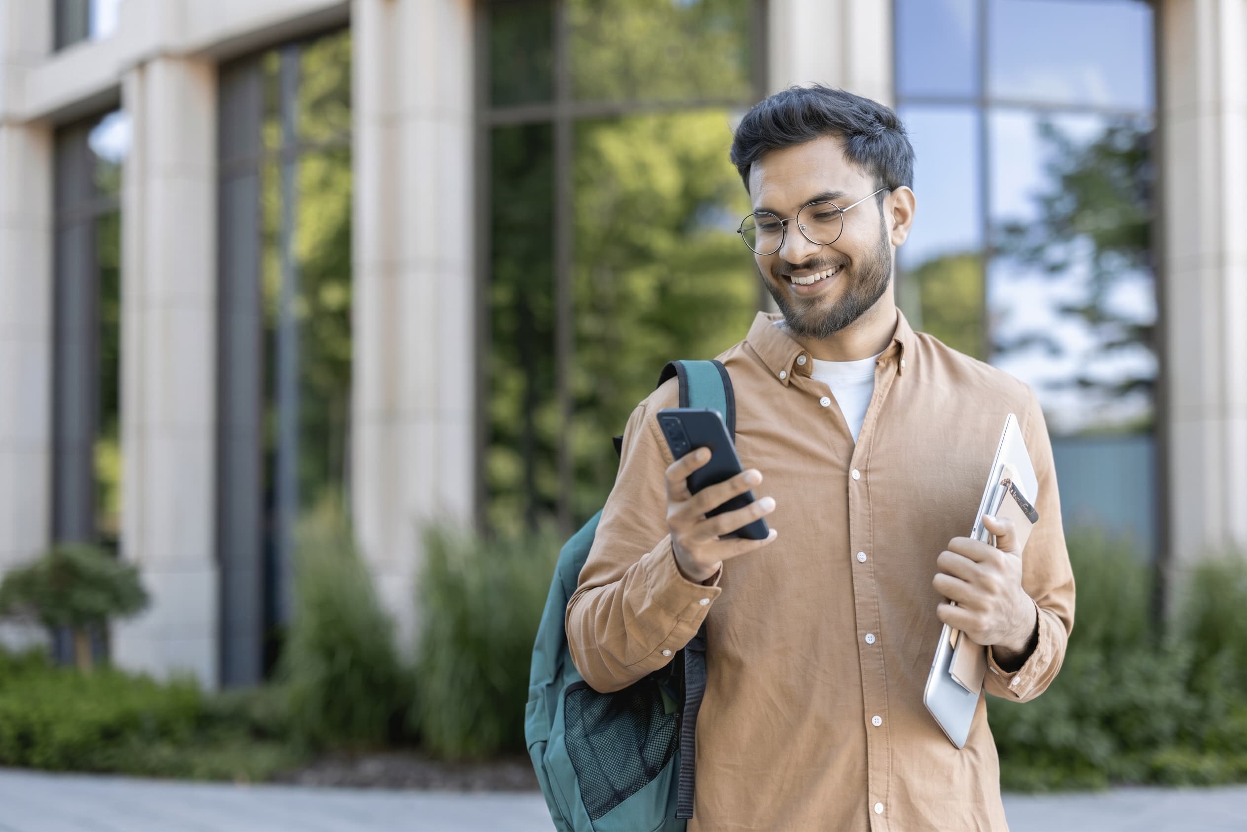 A student outside a modern campus building, smiling at a message on their phone.