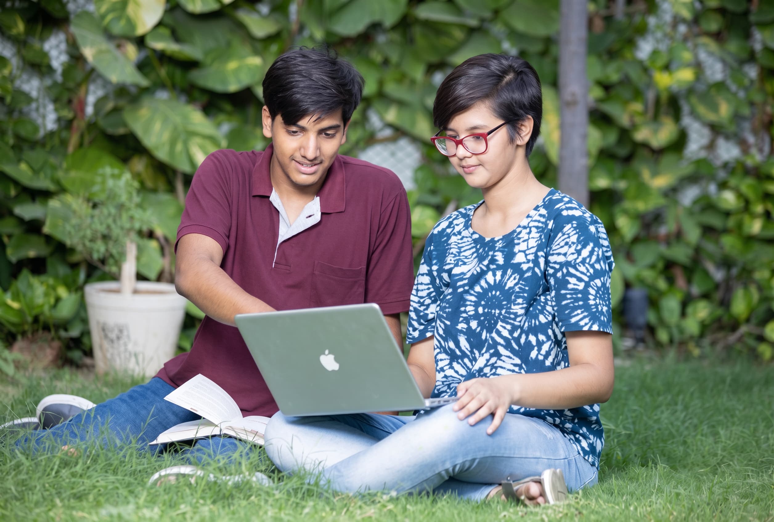 Two teenage students sitting on grass with a laptop and open books.