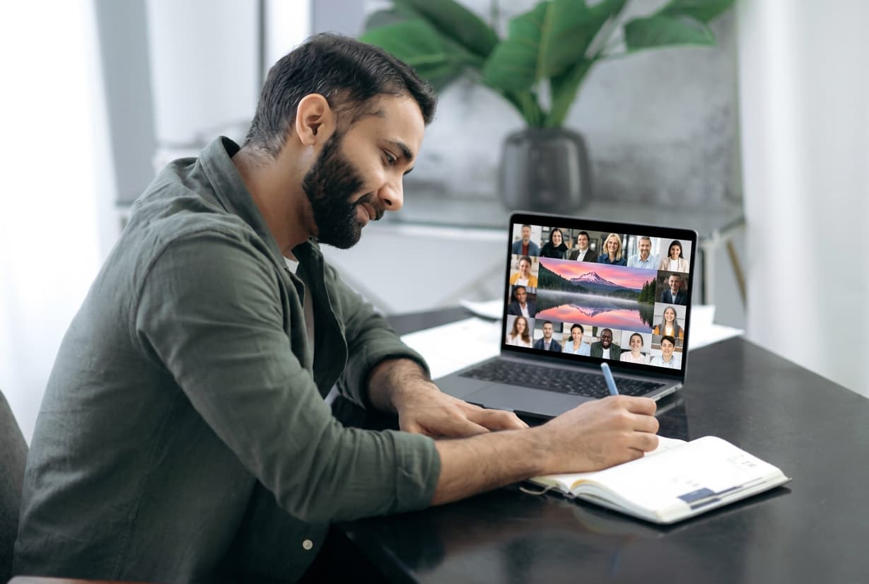 A student on a laptop video call while making notes in a journal.